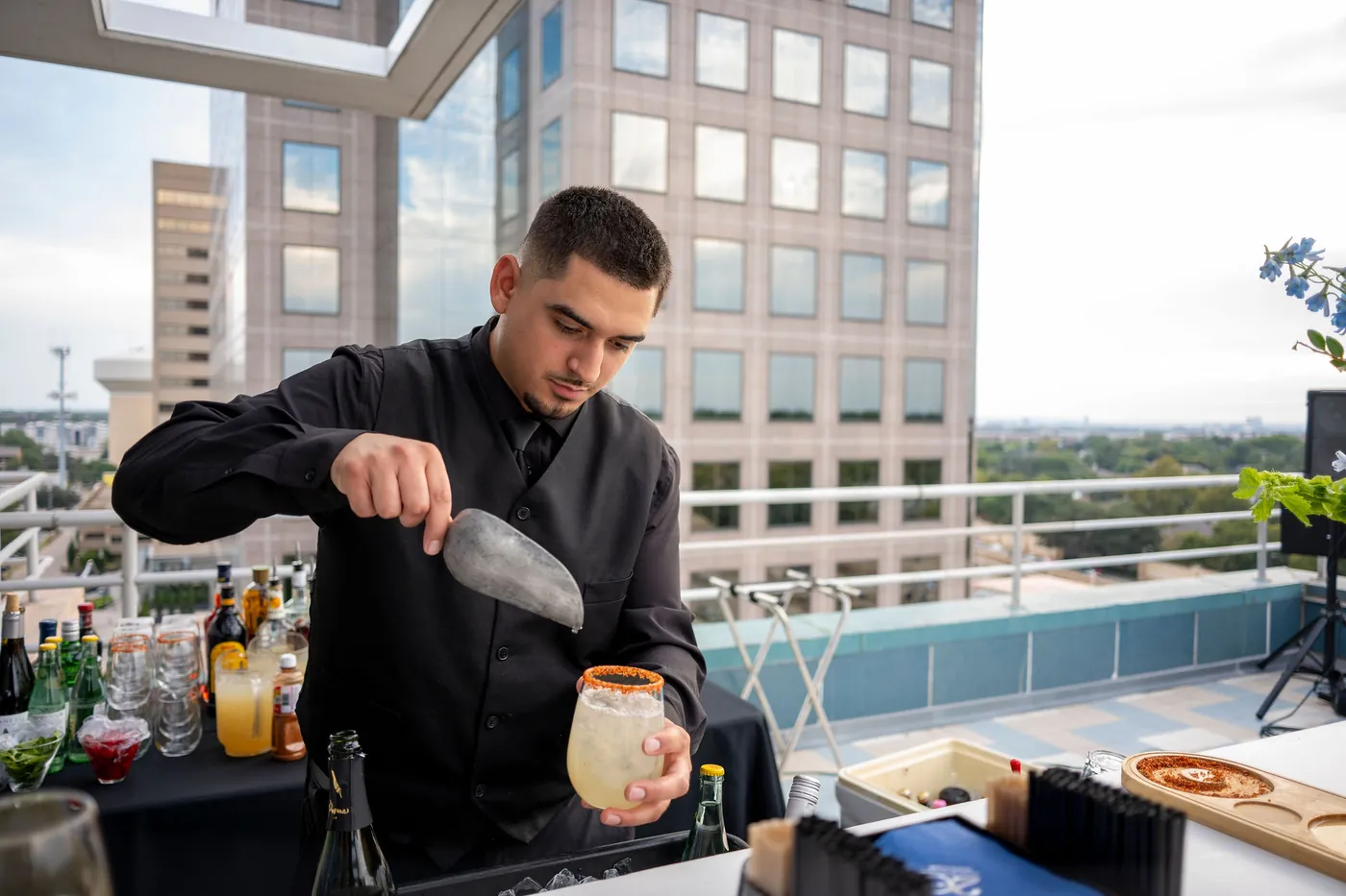 A bartender mixing a drink at a rooftop reception beside a glass tower