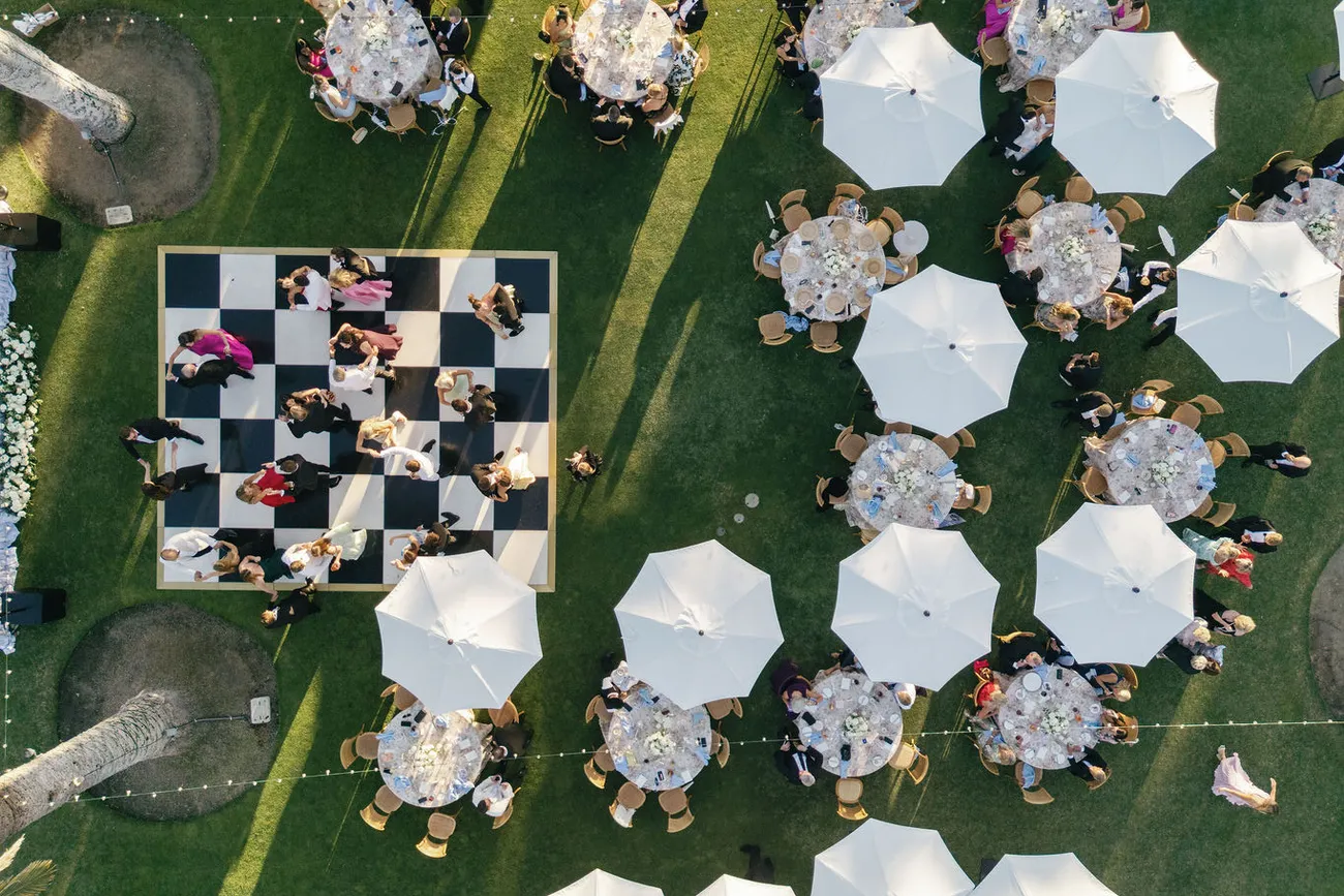 An aerial view of a wedding reception on the lawn, umbrellas set around a checkered dance floor